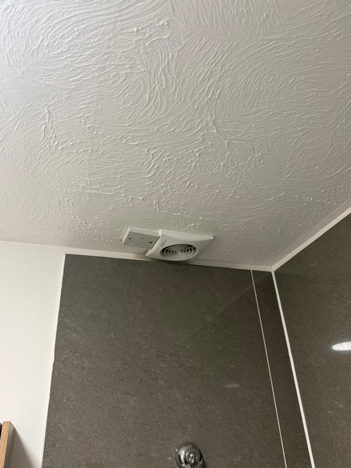 A white extractor fan fitted into a bathroom ceiling above tiled walls.